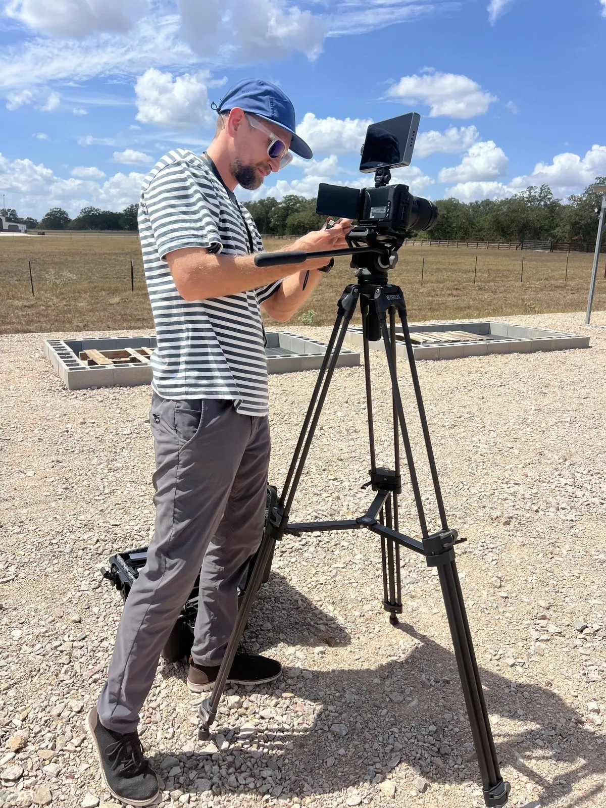 Camera operator setting up a cinema camera on a tripod at the customer's property, concrete forms and pasture behind
