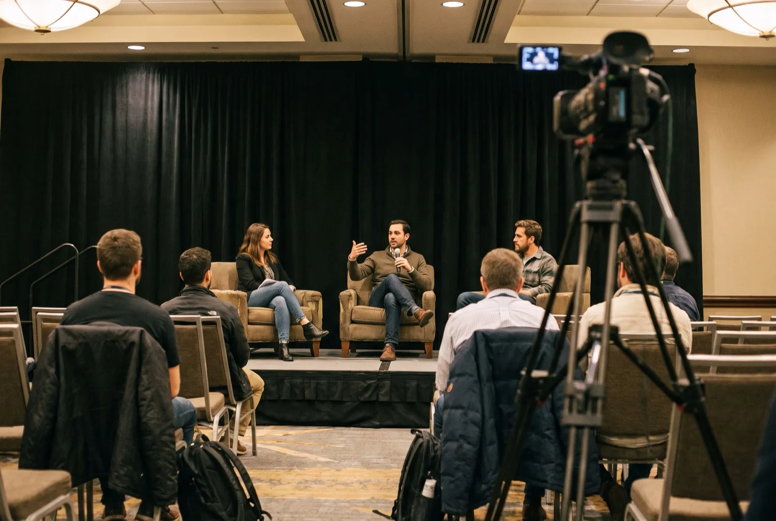 A camera on a tripod records a three-person panel discussion in a hotel breakout room