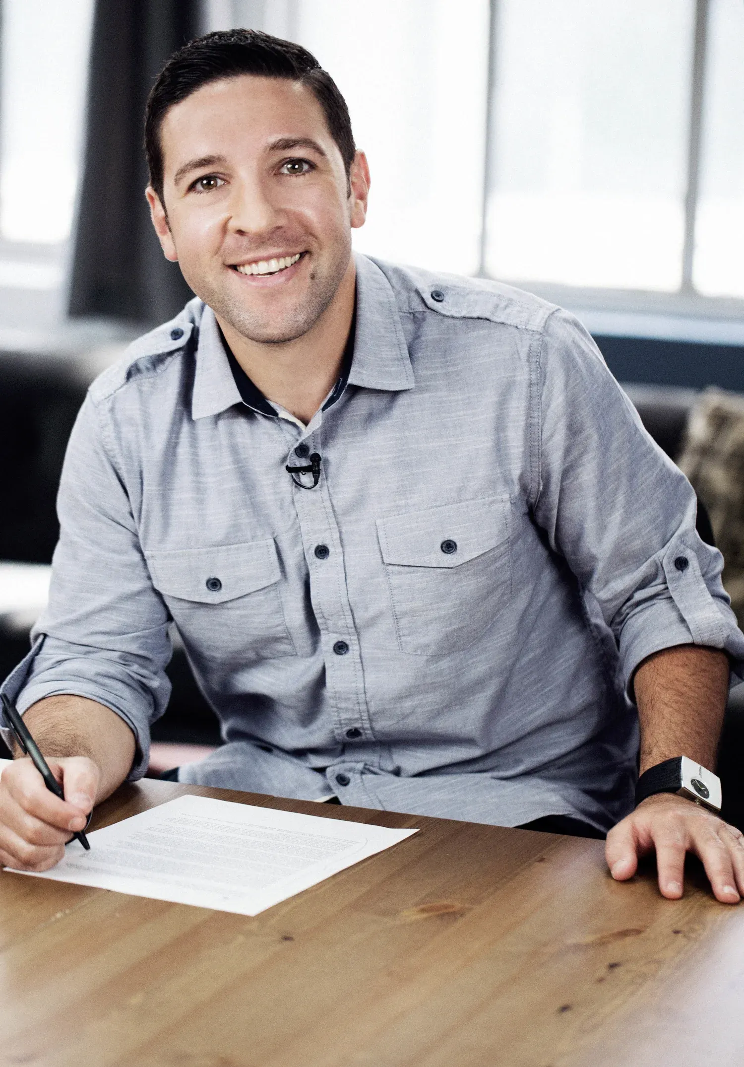 Environmental portrait of a smiling man in a gray shirt writing at a wooden desk in a bright office