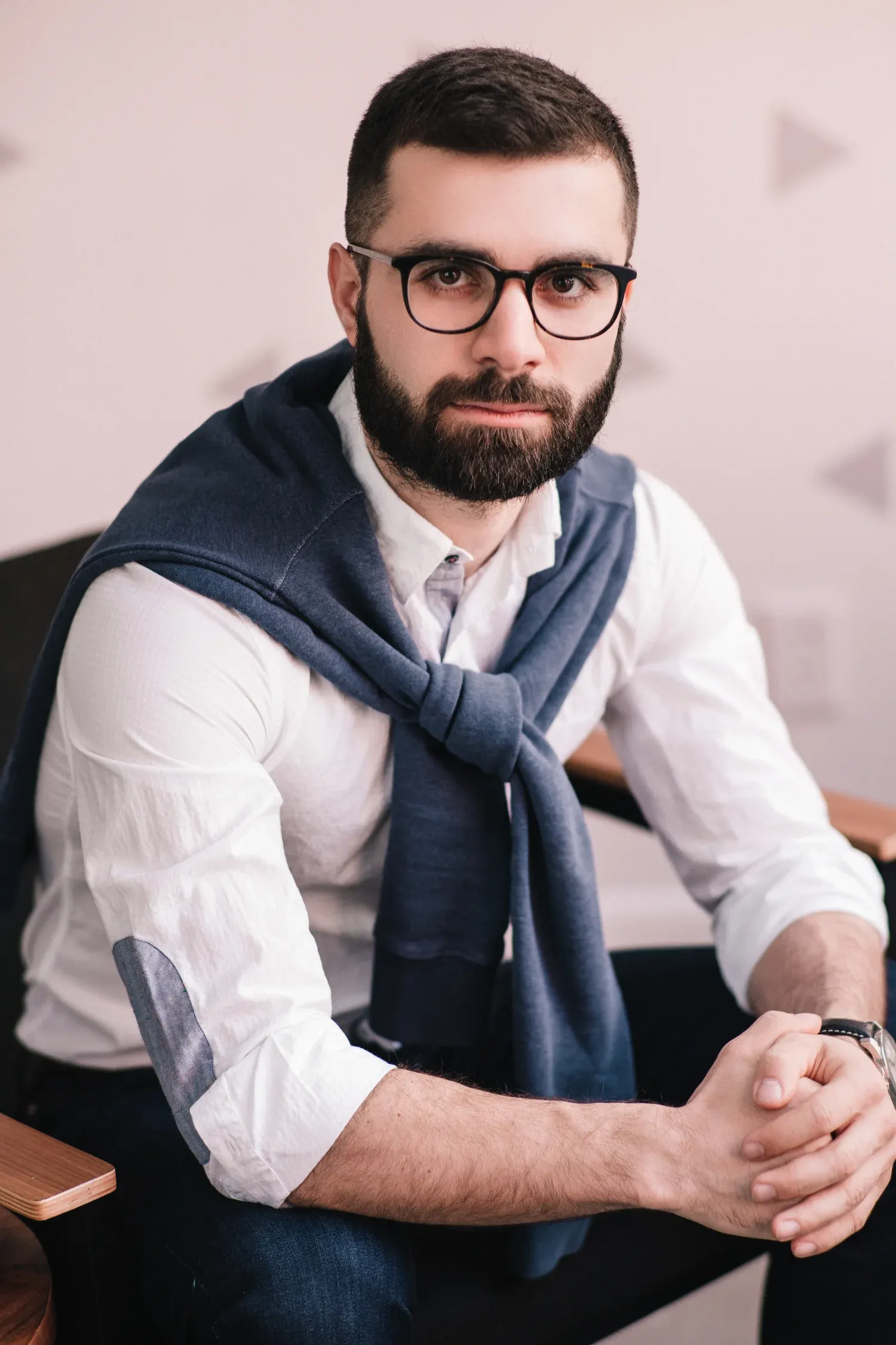 Office portrait of a bearded man in glasses and a white shirt with a sweater over his shoulders, seated in a bright workspace