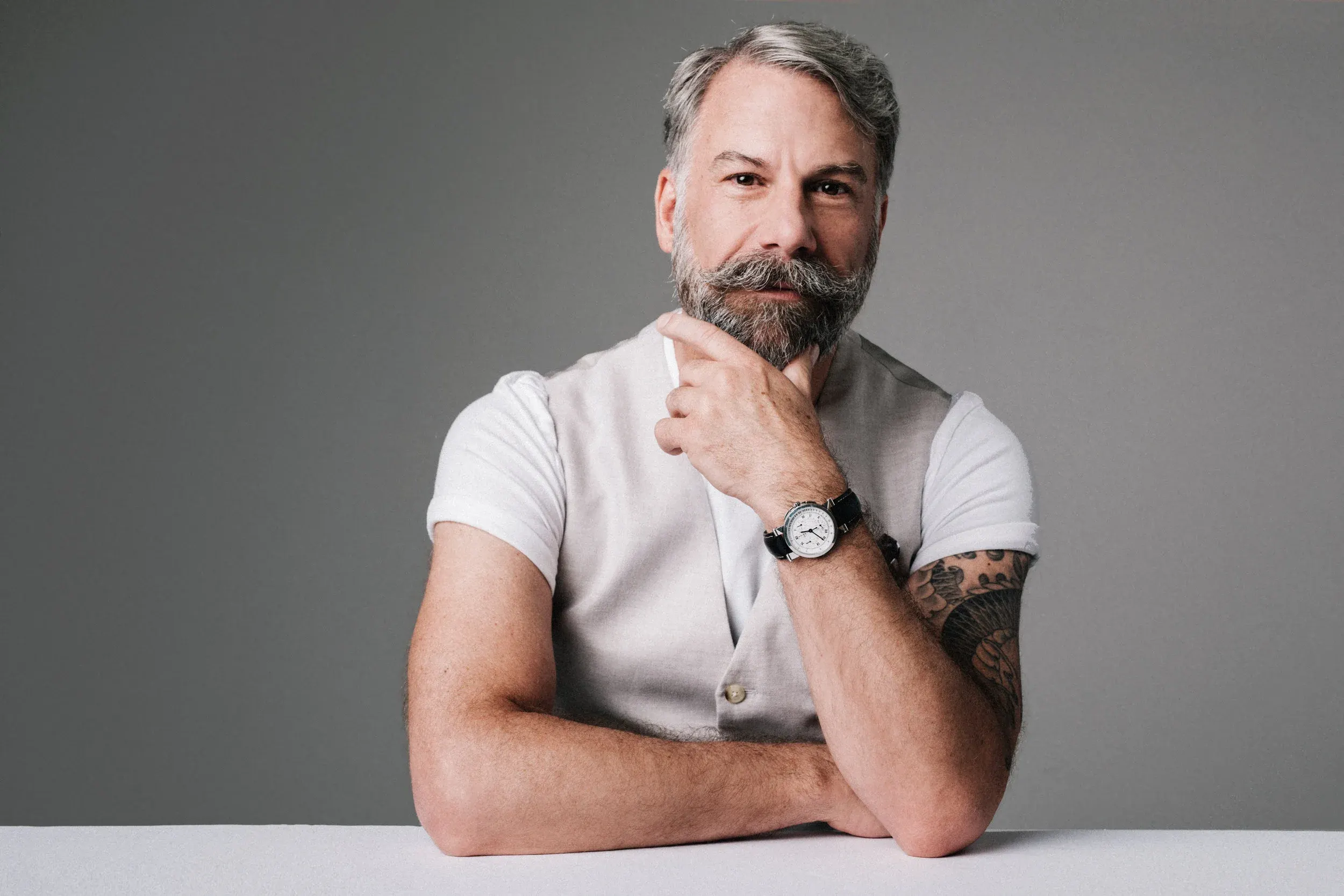 Wide studio portrait of a man with a gray beard in a white tee and vest, hand at his chin, wearing a wristwatch
