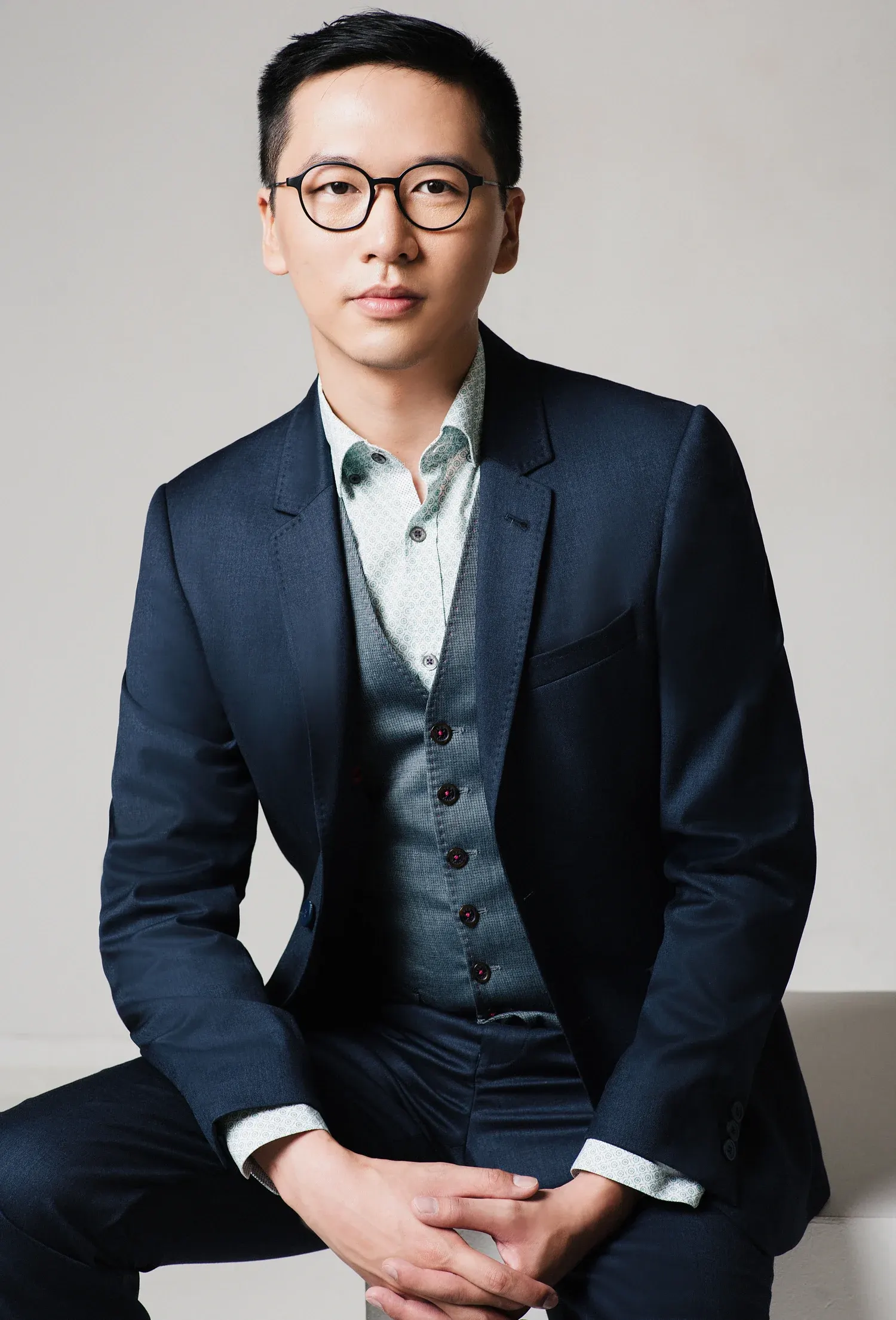 Corporate headshot of a man in glasses and a navy three-piece suit, seated against a white studio backdrop