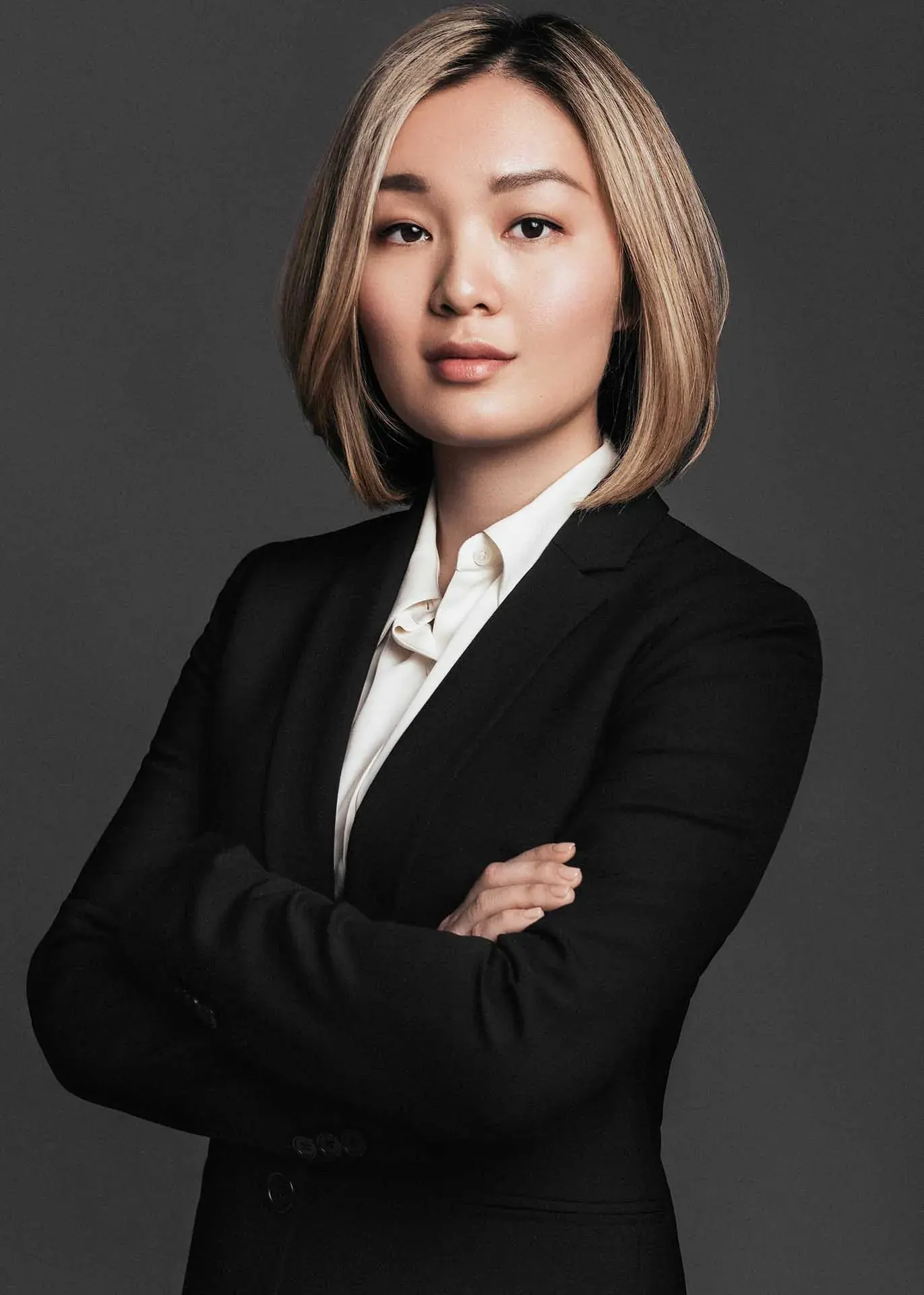 Executive headshot of a woman with a dark bob in a black blazer and white shirt, arms crossed, on a dark gray backdrop