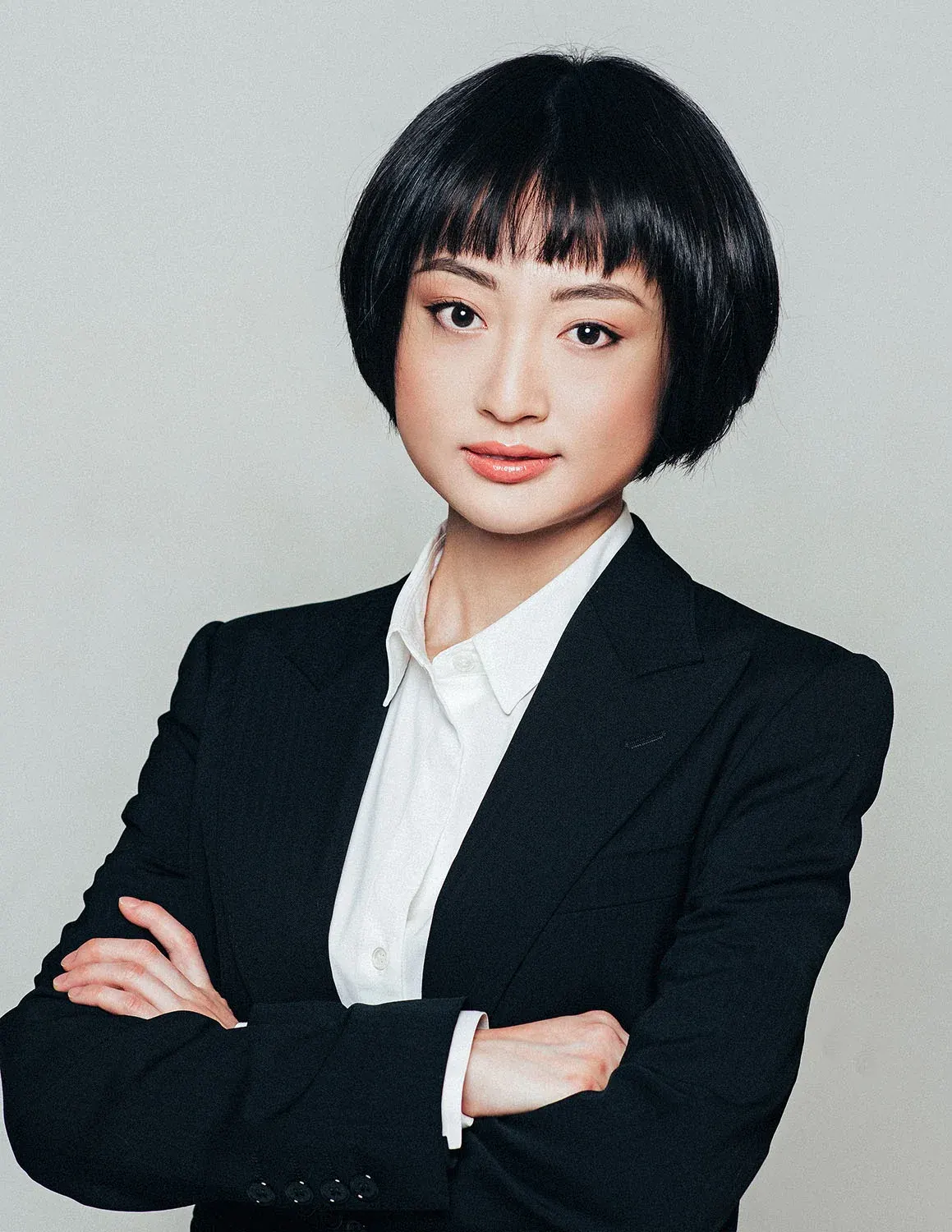 Professional headshot of a woman with a dark bob in a black suit and white shirt, arms crossed, on a gray backdrop