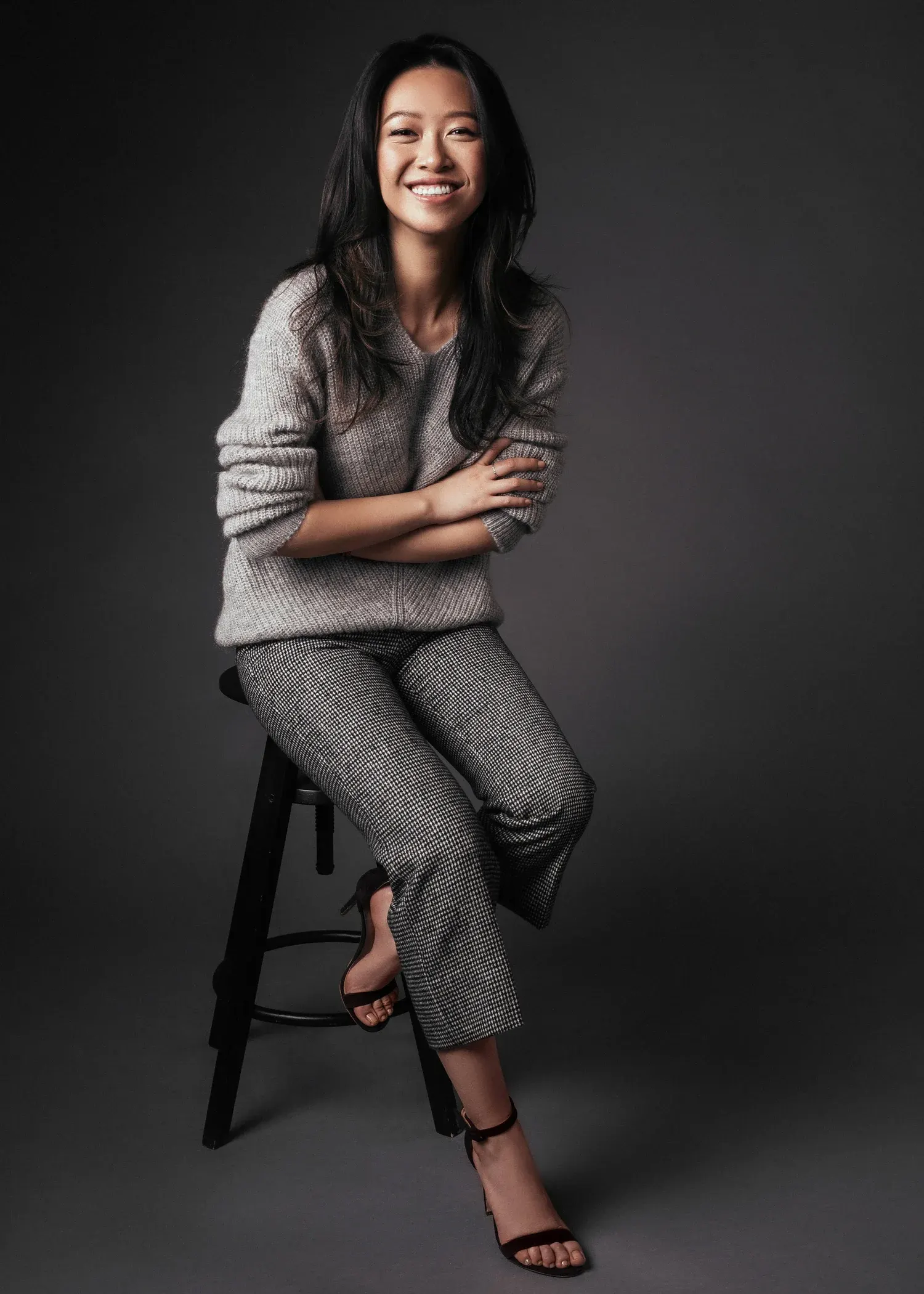 Full-length studio portrait of a laughing woman in a gray sweater and trousers seated on a stool against a dark backdrop