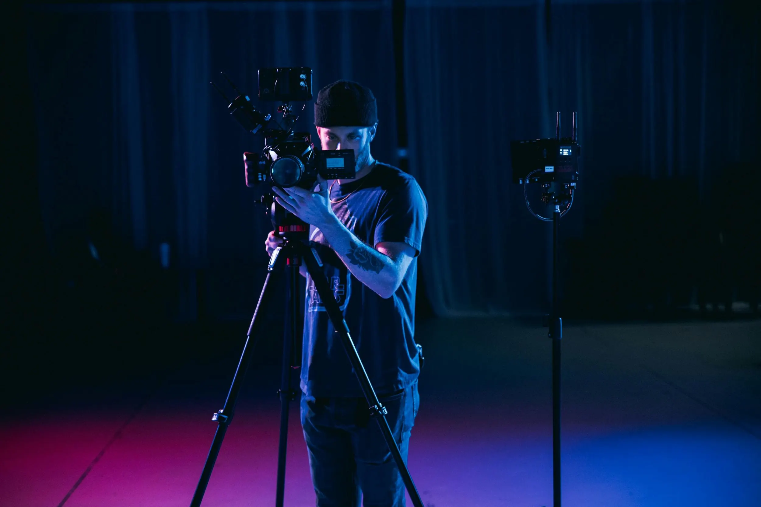 Camera operator framing a shot on a cinema camera in a studio lit with blue and pink light